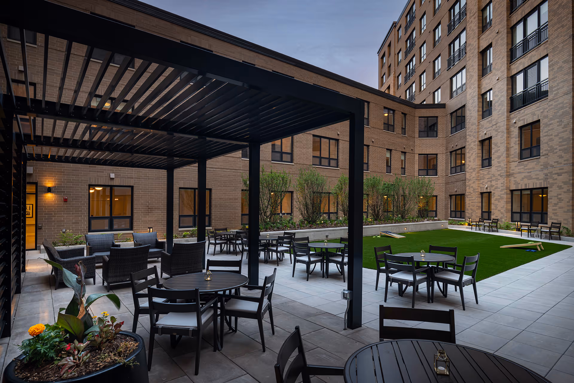 Outdoor courtyard with a pergola-covered seating area, round tables, artificial lawn and cornhole boards surrounded by the apartment building.