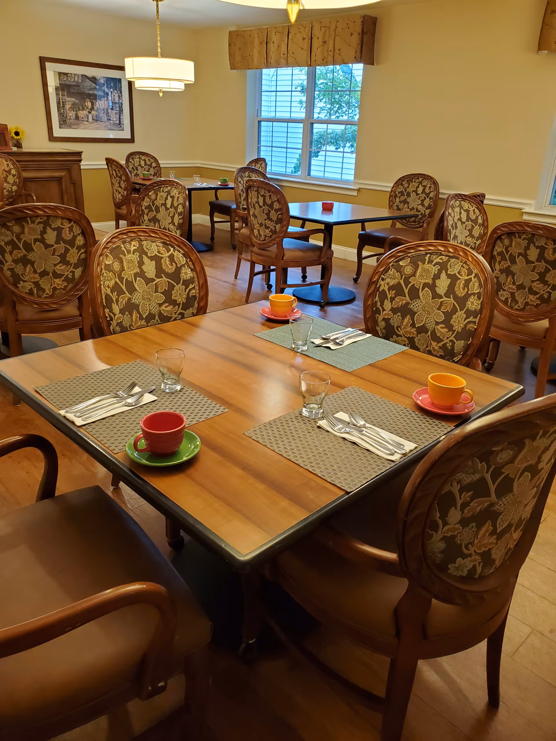 A dining room with wooden tables and floral upholstered chairs. Each table is set with placemats, glasses, silverware, and colorful cups and saucers. There is a window with a valance letting in natural light, and a framed picture on the wall.