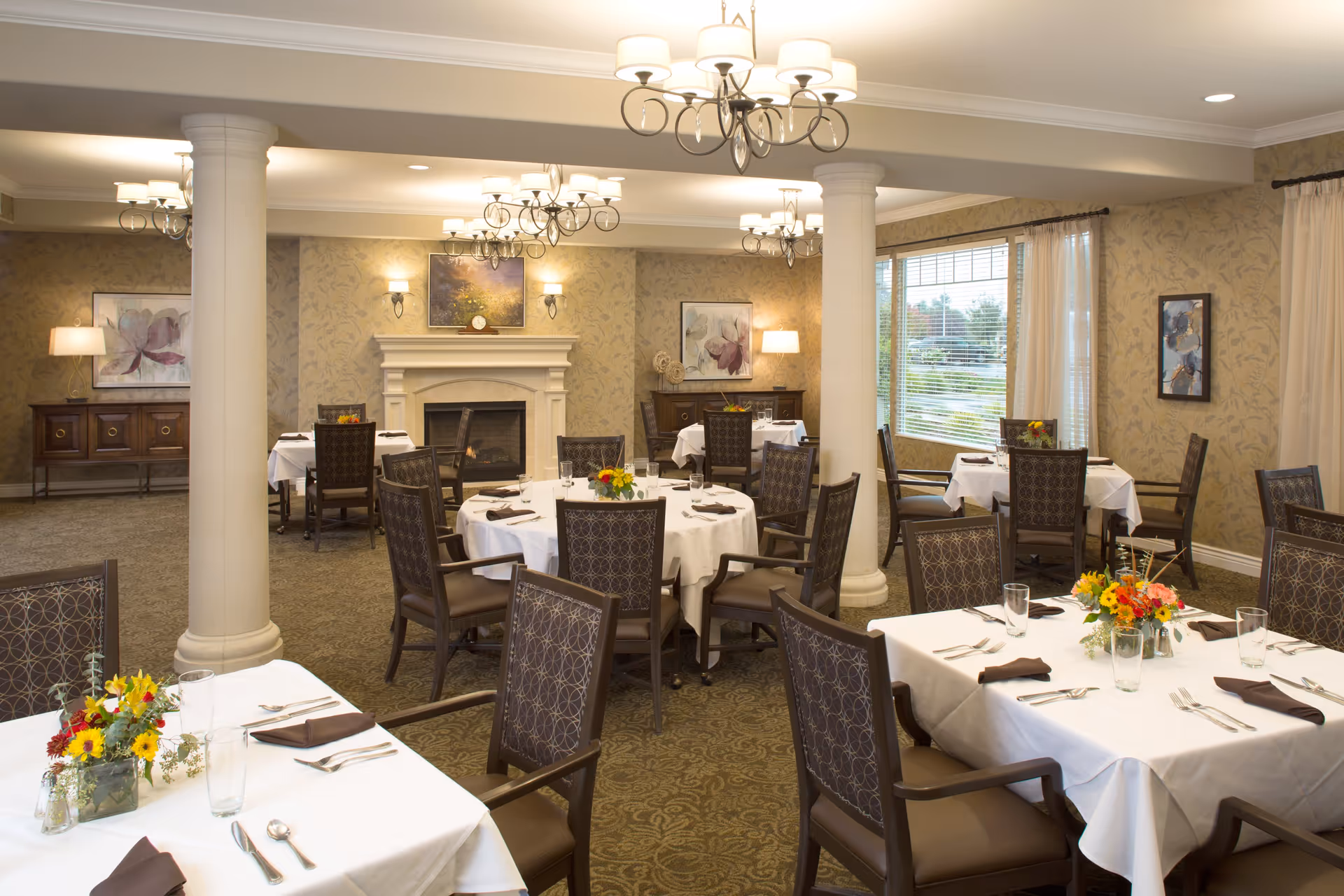 A well-lit dining room with round and square tables covered with white tablecloths, each set with brown napkins, glasses, and silverware. The room features patterned carpet, beige floral wallpaper, large windows with curtains, decorative columns, chandeliers, and a fireplace with artwork above it. Flower arrangements are placed on the tables.