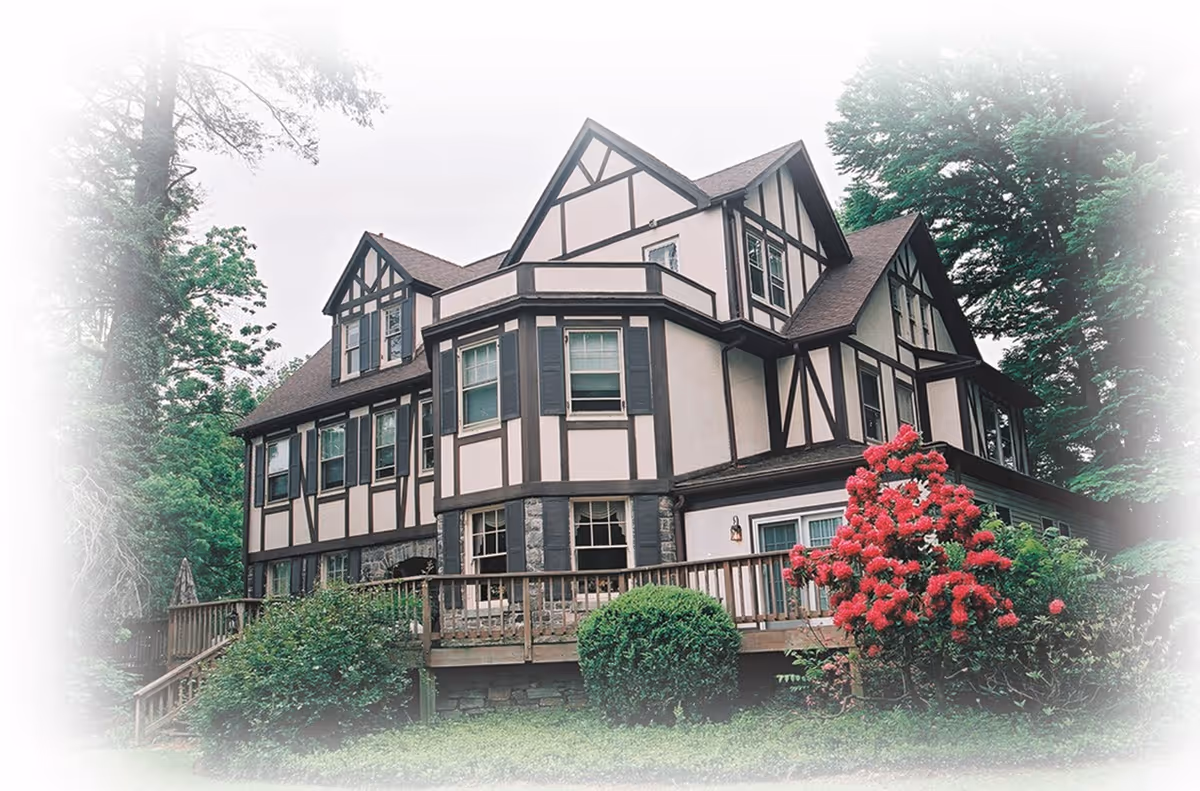 Tudor-style multi-story house with a wooden deck, surrounded by trees and flowering bushes.