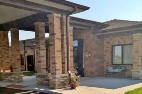 Brick nursing facility entrance with columns, a covered walkway, and a bench outside.