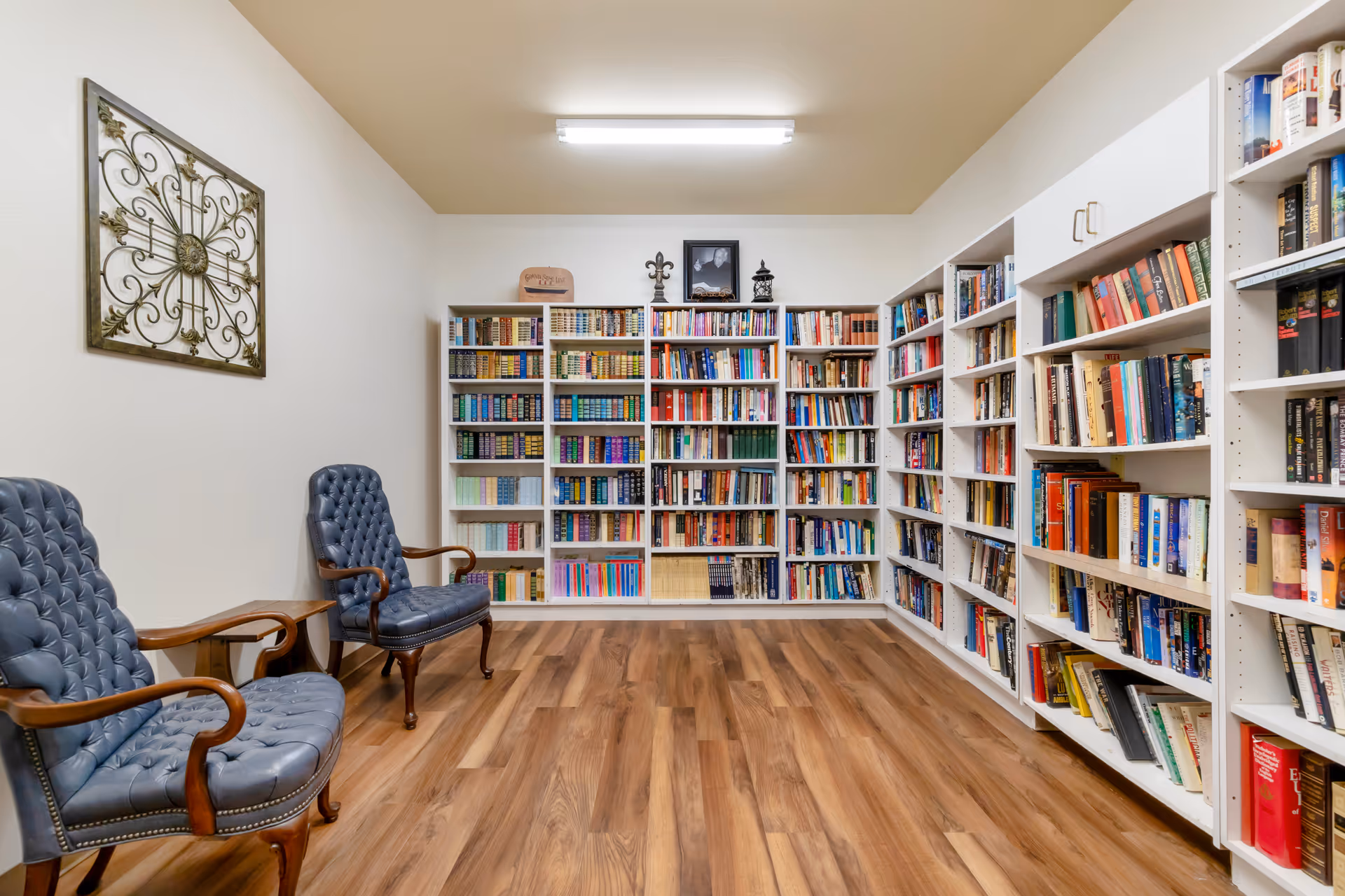 A small library room with white bookshelves filled with books, two blue upholstered armchairs, and wood flooring.