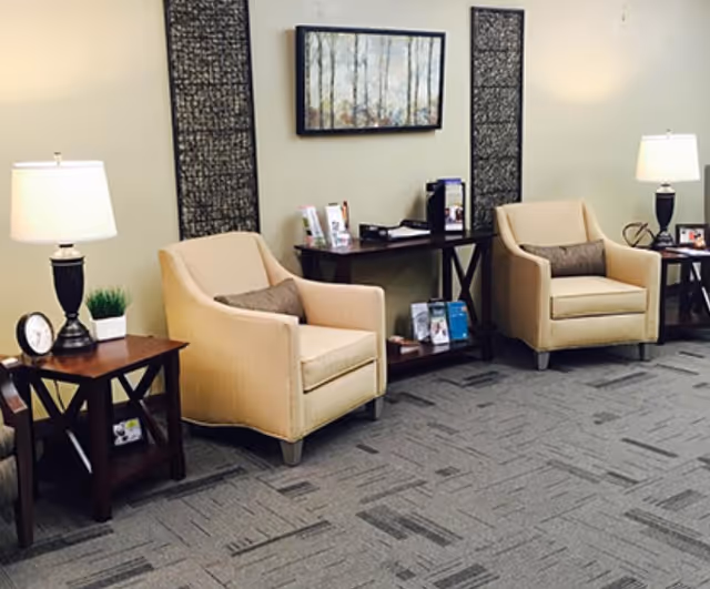 Two beige armchairs with side tables and lamps arranged in a carpeted lounge area with wall art and a console table.