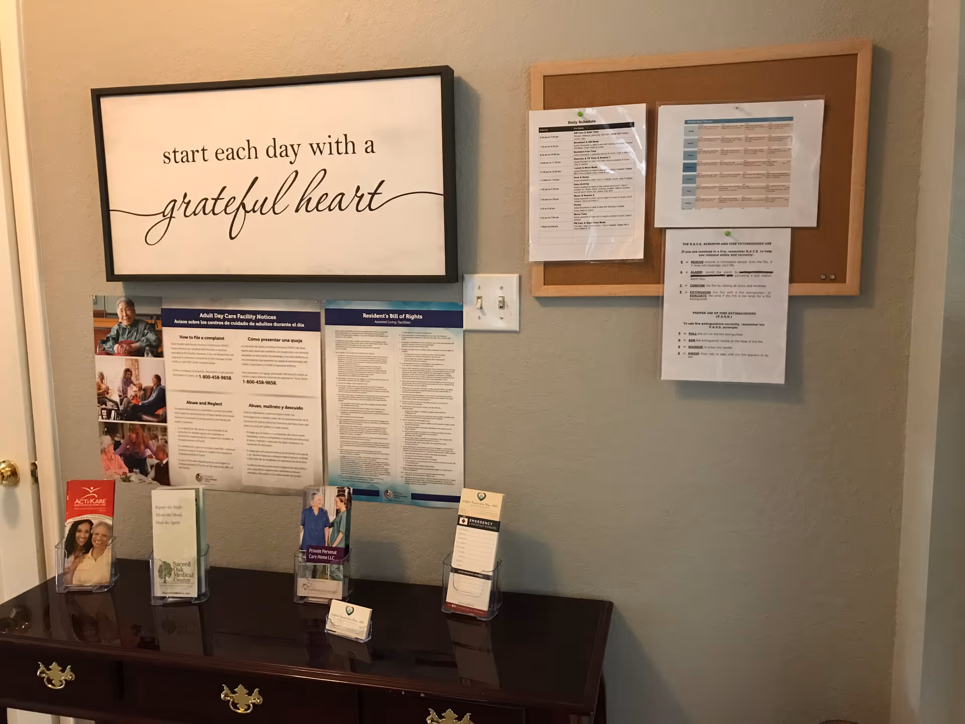 Interior wall with a framed sign reading 'start each day with a grateful heart', bulletin boards, and brochures on a wooden console table.