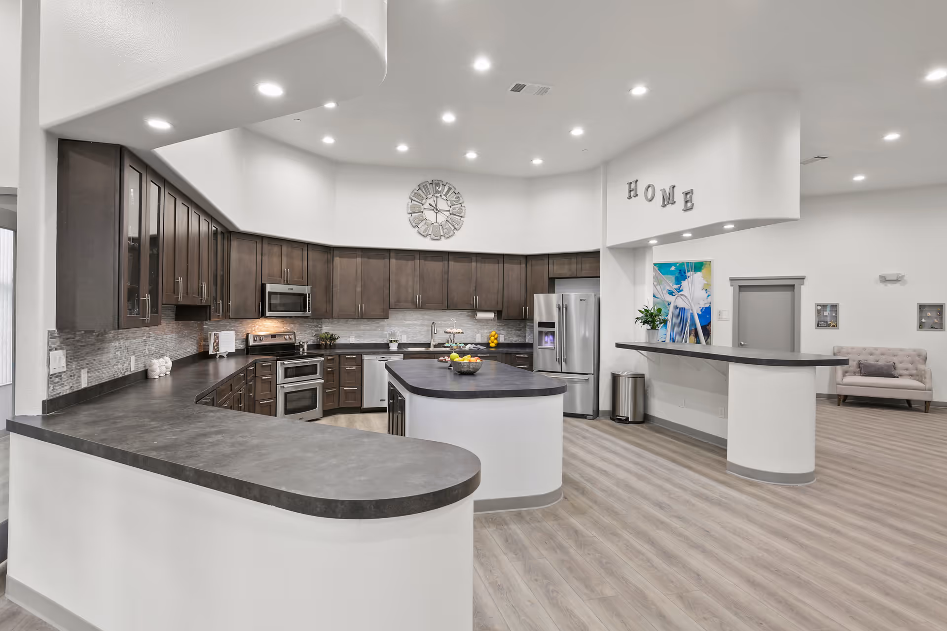 Modern kitchen area with dark wood cabinets, stainless steel appliances including a refrigerator, microwave, and oven. The kitchen has a large island with a bowl of fruit on it, light wood flooring, and bright recessed ceiling lights. The word 'HOME' is displayed on the wall above a counter with a colorful painting and a small plant. In the background, there is a seating area with a cushioned bench.