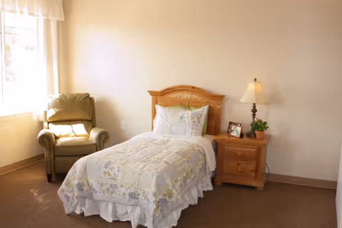 A cozy bedroom in an assisted living facility featuring a single bed with a floral quilt and pillows, a wooden nightstand with a lamp, a small framed photo, and a potted plant. There is a comfortable armchair near a window with sheer curtains allowing natural light to fill the room.