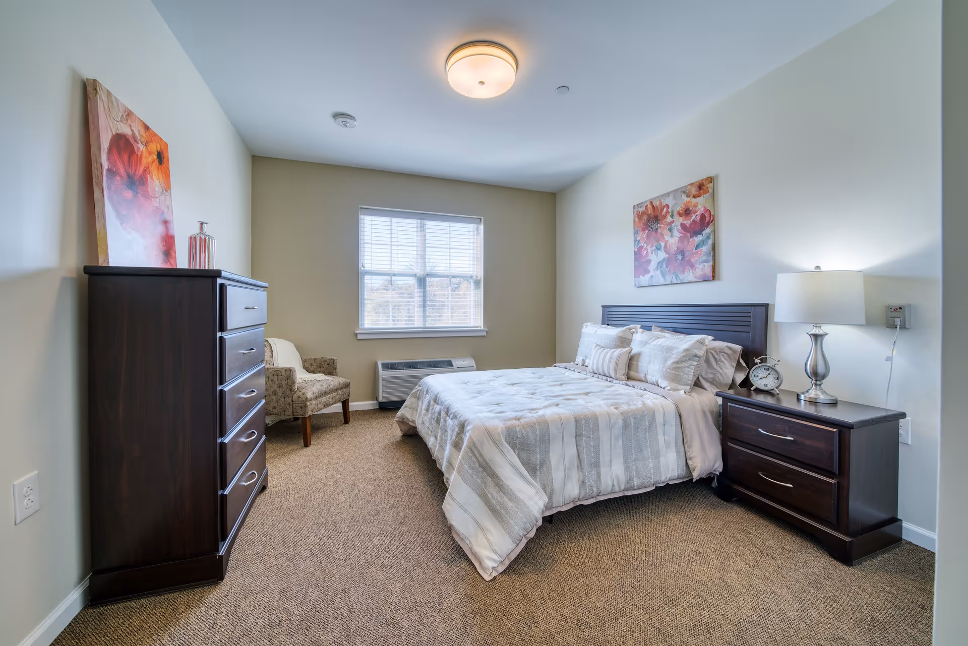 Well-lit furnished bedroom with a bed, nightstand, dresser, chair, and a window.