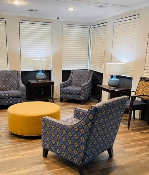A cozy sitting area with four patterned armchairs arranged around a large round yellow ottoman. Two wooden side tables with blue ceramic lamps are placed near the chairs. The room has multiple windows with white blinds and wood flooring.