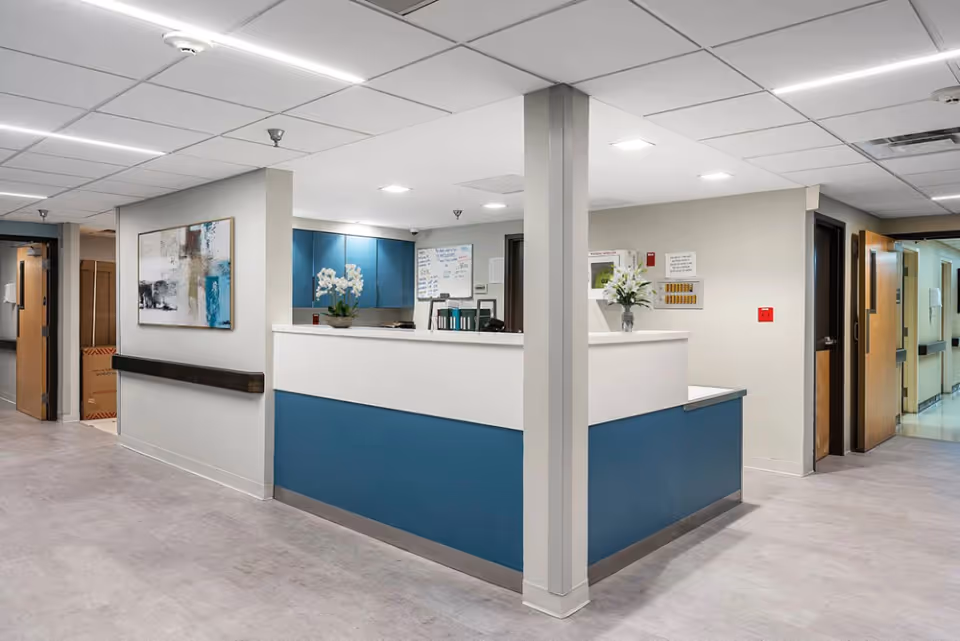 Interior view of a nursing center reception area with a blue and white reception desk, a whiteboard on the wall, a potted plant with white flowers on the desk, and hallways leading to other rooms.