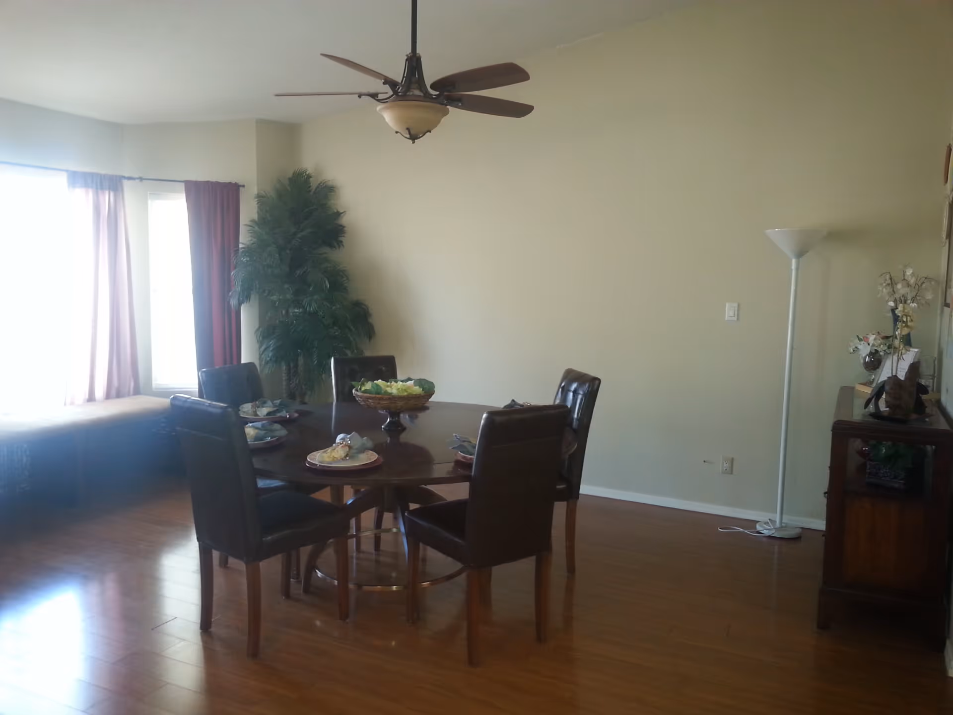 A dining area with a round wooden table set with plates and napkins, surrounded by six dark brown leather chairs. There is a ceiling fan with lights above the table. To the left, there is a window with curtains and a cushioned bench underneath. A tall green plant is placed in the corner near the window. On the right side, there is a white floor lamp and a wooden cabinet with decorative items on top. The room has wooden flooring and light-colored walls.