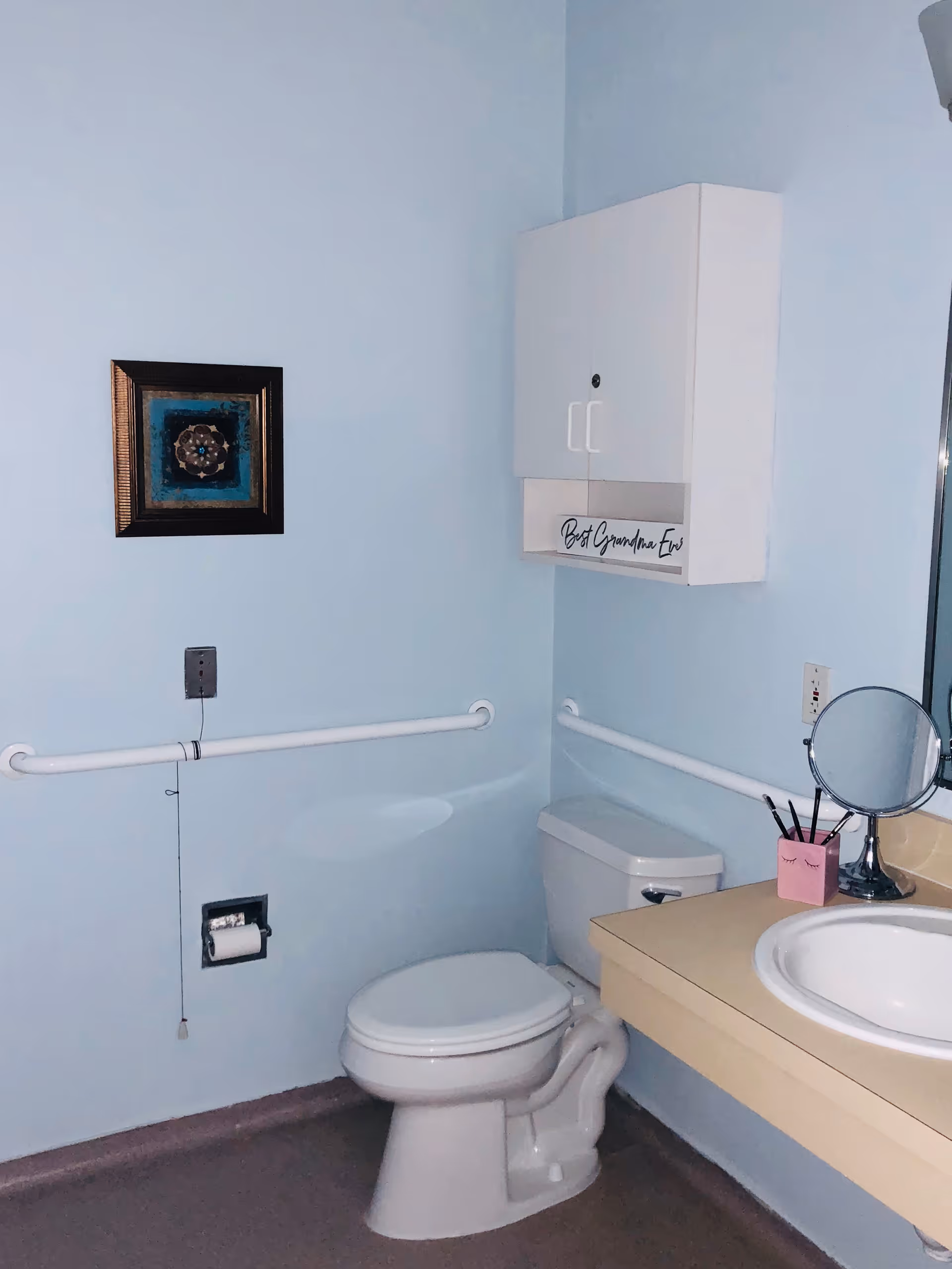 A small bathroom with light blue walls featuring a white toilet, a beige countertop with a white sink, a round mirror, and a pink container holding makeup brushes. There are white grab bars on the walls for support, a white wall-mounted cabinet with the text 'Best Grandma Ever' on the bottom shelf, and a framed decorative artwork hanging on the wall.