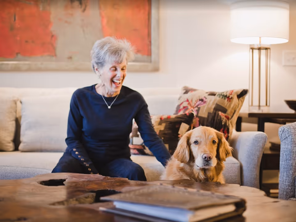 An older woman sitting on a sofa laughing while petting a golden retriever in a cozy living room.