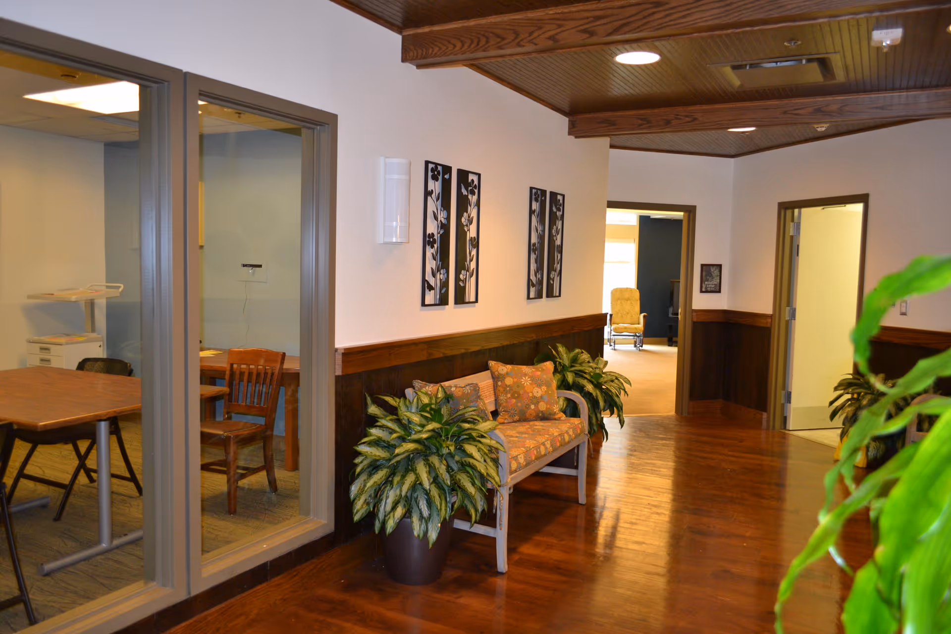 Interior hallway of a senior living facility with wooden floors and ceiling. There is a bench with floral cushions and two potted plants on either side. On the left, there is a glass-walled room with a wooden table and chairs. The hallway leads to two open doorways, one showing a room with a yellow armchair and the other a room with a white door. The walls are decorated with three black and white floral art pieces.