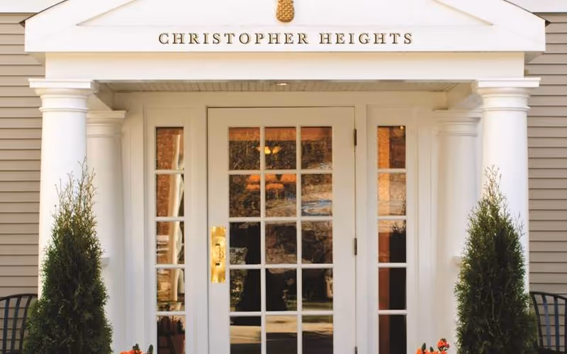 Entrance of Christopher Heights facility with white double doors featuring multiple glass panes, flanked by two white columns and two tall green shrubs. The facility name 'CHRISTOPHER HEIGHTS' is displayed above the entrance.