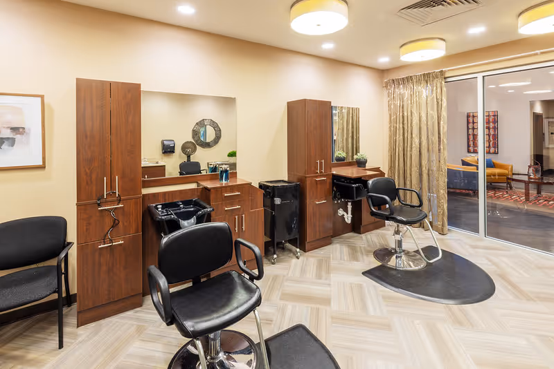 Interior view of a hair salon area in a senior living facility with two black salon chairs in front of wooden cabinets and sinks for hair washing. There is a large mirror on the wall, a black waiting chair to the left, and a glass sliding door with gold curtains leading to another room with seating visible.