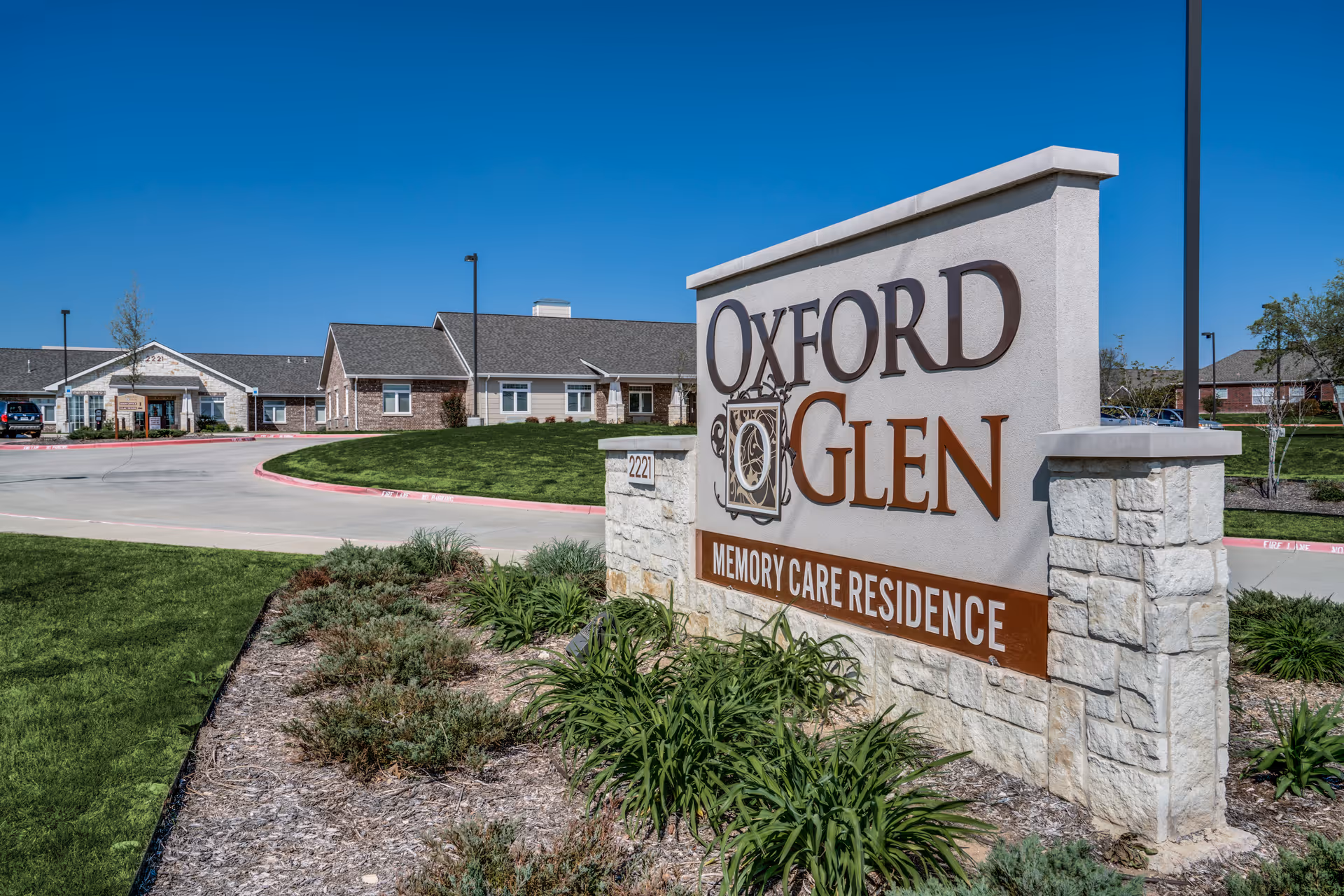 Outdoor view of Oxford Glen Memory Care Residence building with a large stone sign in the foreground displaying the facility name. The building is single-story with a combination of brick and siding, surrounded by green grass and landscaping under a clear blue sky.