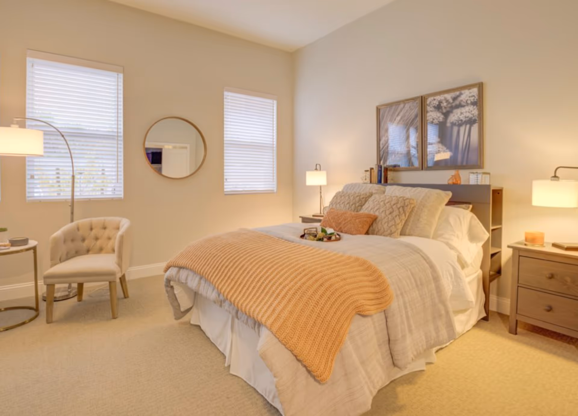 Sunlit, neatly made bedroom with a bed dressed in neutral linens and a peach throw, bedside tables with lamps, a tufted chair, and two windows.
