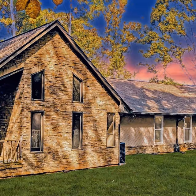 Exterior view of a senior living facility building with stone and wood siding, multiple windows, and a sloped roof, set against a backdrop of trees with autumn foliage and a colorful sunset sky.