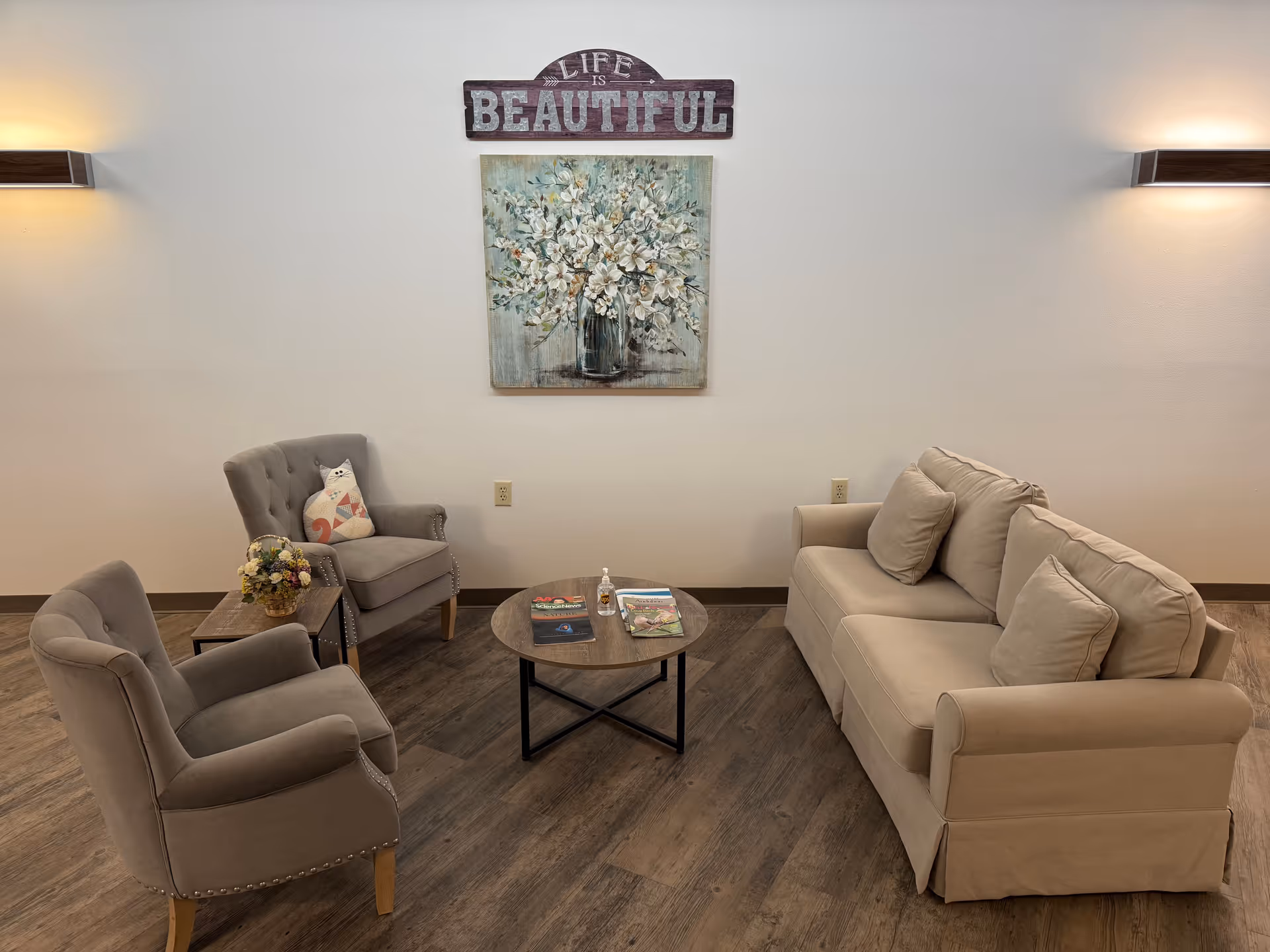 A cozy seating area with a beige sofa, two gray armchairs, a round wooden coffee table with magazines and hand sanitizer, and a small side table with a flower arrangement. On the wall above is a painting of white flowers in a vase and a wooden sign that reads 'Life is Beautiful'.