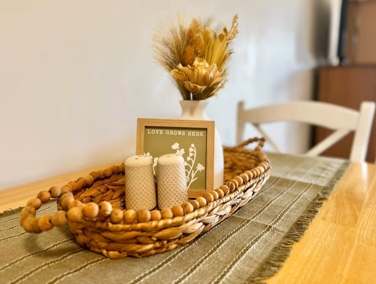 A woven basket with wooden bead handles sits on a green table runner on a wooden table. Inside the basket are two white textured salt and pepper shakers, a small framed sign that reads 'LOVE GROWS HERE,' and a white vase holding dried flowers and grasses. A white chair is visible in the background.
