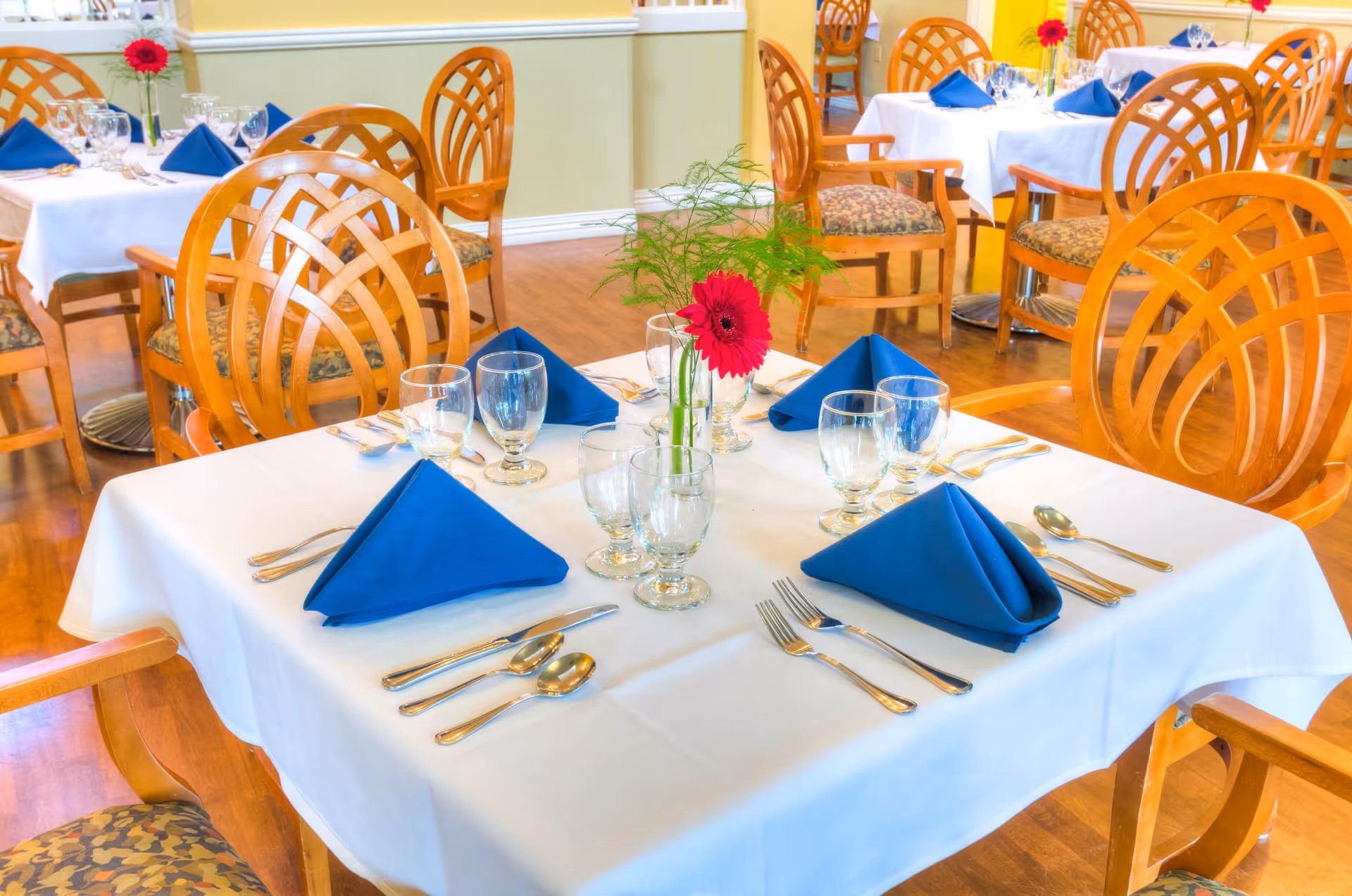 A dining room table set for four with white tablecloth, blue folded napkins, multiple glasses, silverware, and a small vase with a red flower and greenery. The room has wooden chairs with patterned cushions and a polished wooden floor.