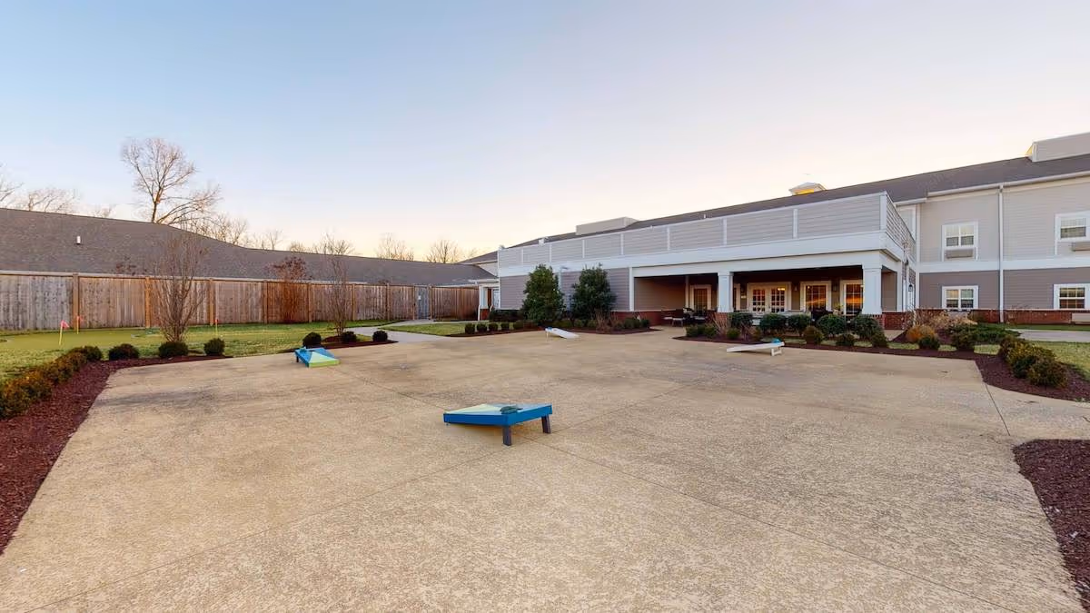 Outdoor courtyard area at The Goldton at Spring Hill featuring a concrete patio with three cornhole game boards, surrounded by landscaped bushes and trees, with a two-story building in the background under a clear sky.