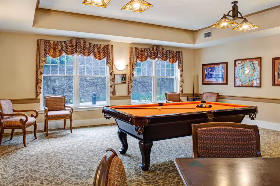 A bright recreation room with an orange-felt pool table, upholstered chairs, framed artwork, and large windows with draped valances.