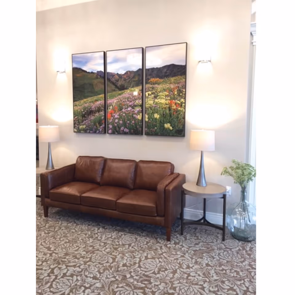 A brown leather sofa sits against a beige wall with a three-panel landscape painting of a flower field and mountains above it. On either side of the sofa are small round tables with modern lamps. To the right, there is a large glass vase with green foliage. The floor is covered with a patterned carpet.