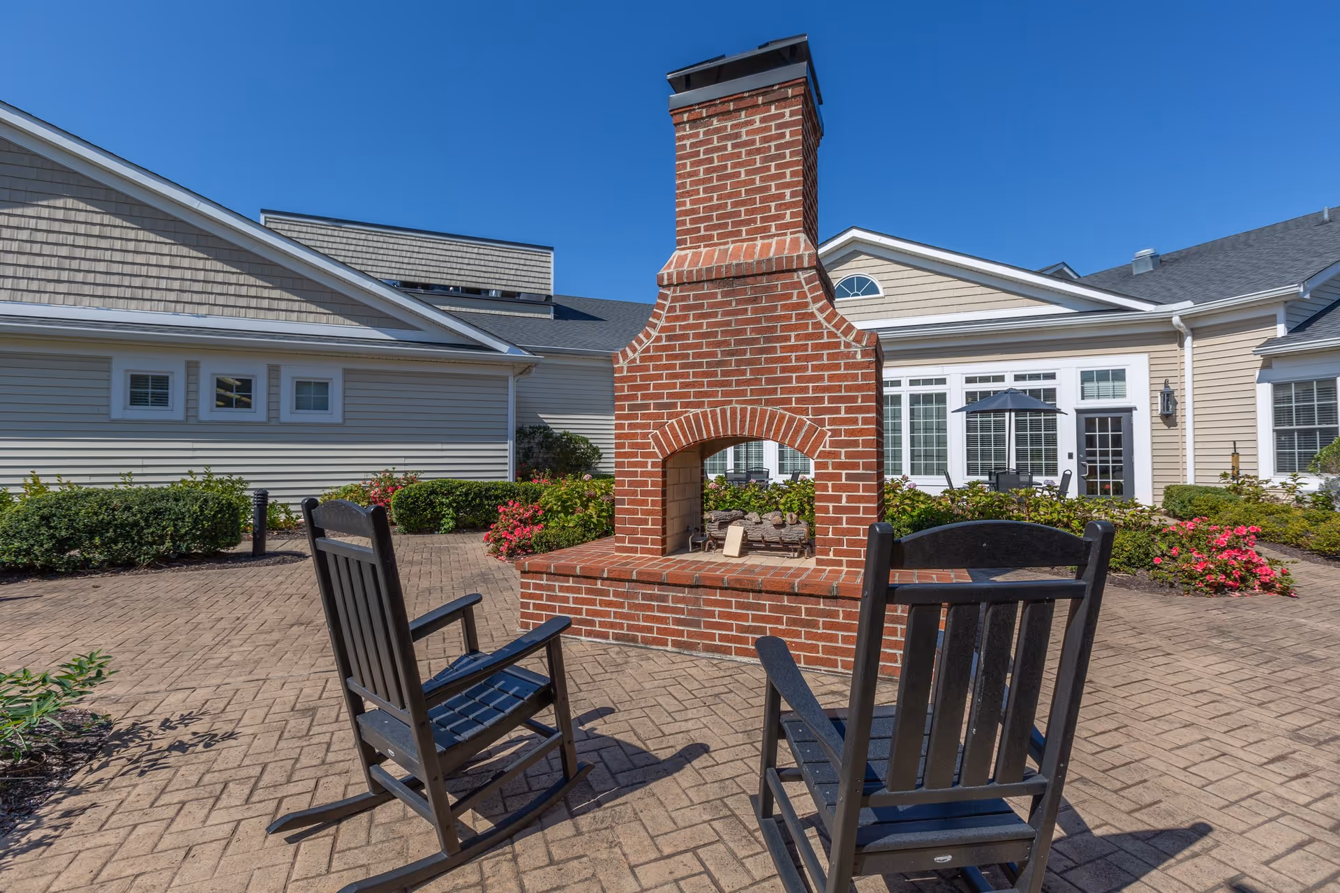 Brick outdoor fireplace with two wooden rocking chairs on a paved courtyard in front of the senior living building.