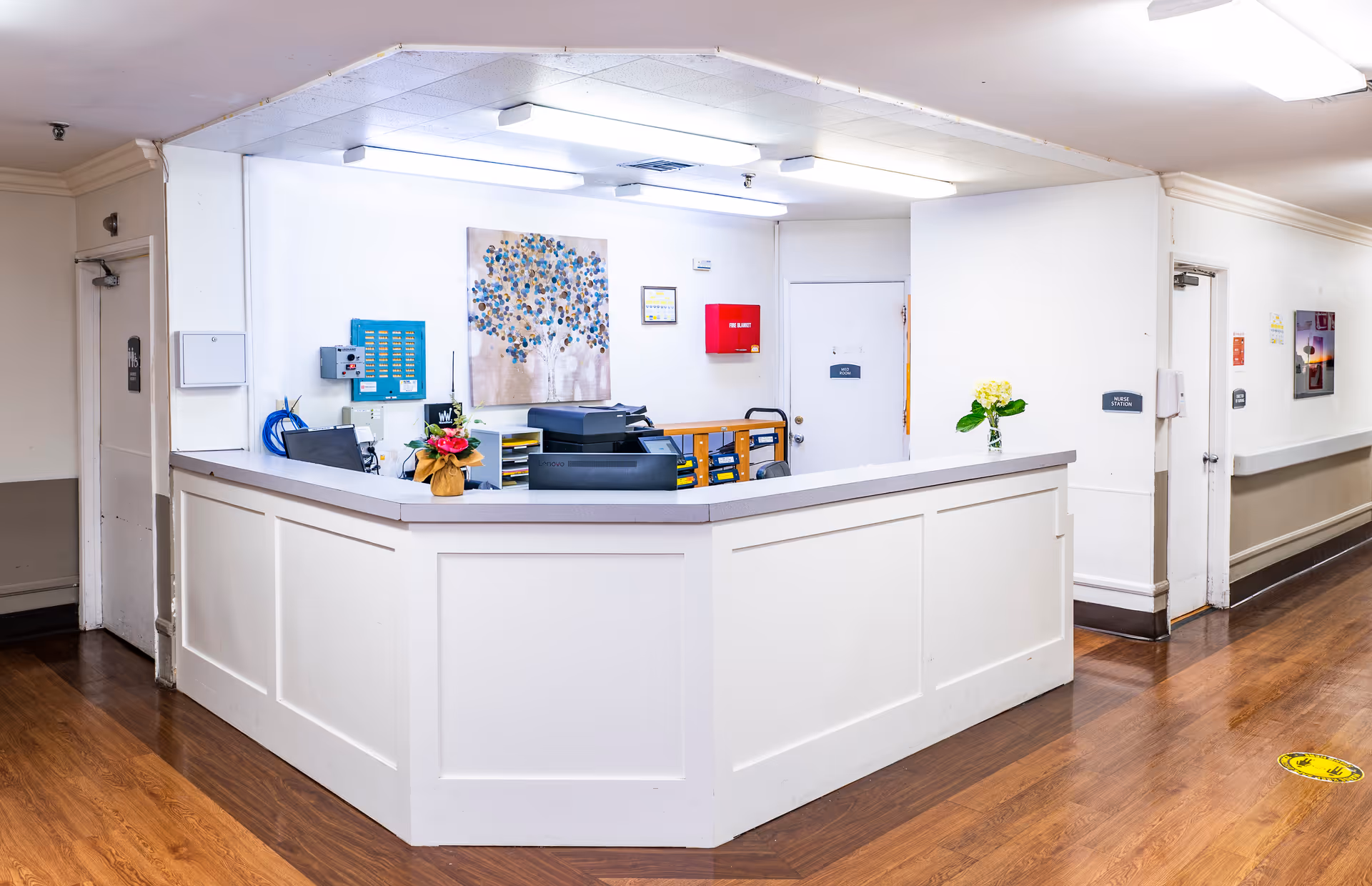 Reception desk area inside a senior living facility with a white paneled counter, office equipment, and small flower arrangements on the counter. The walls are white with a colorful tree painting, and there are doors and hallway visible in the background.