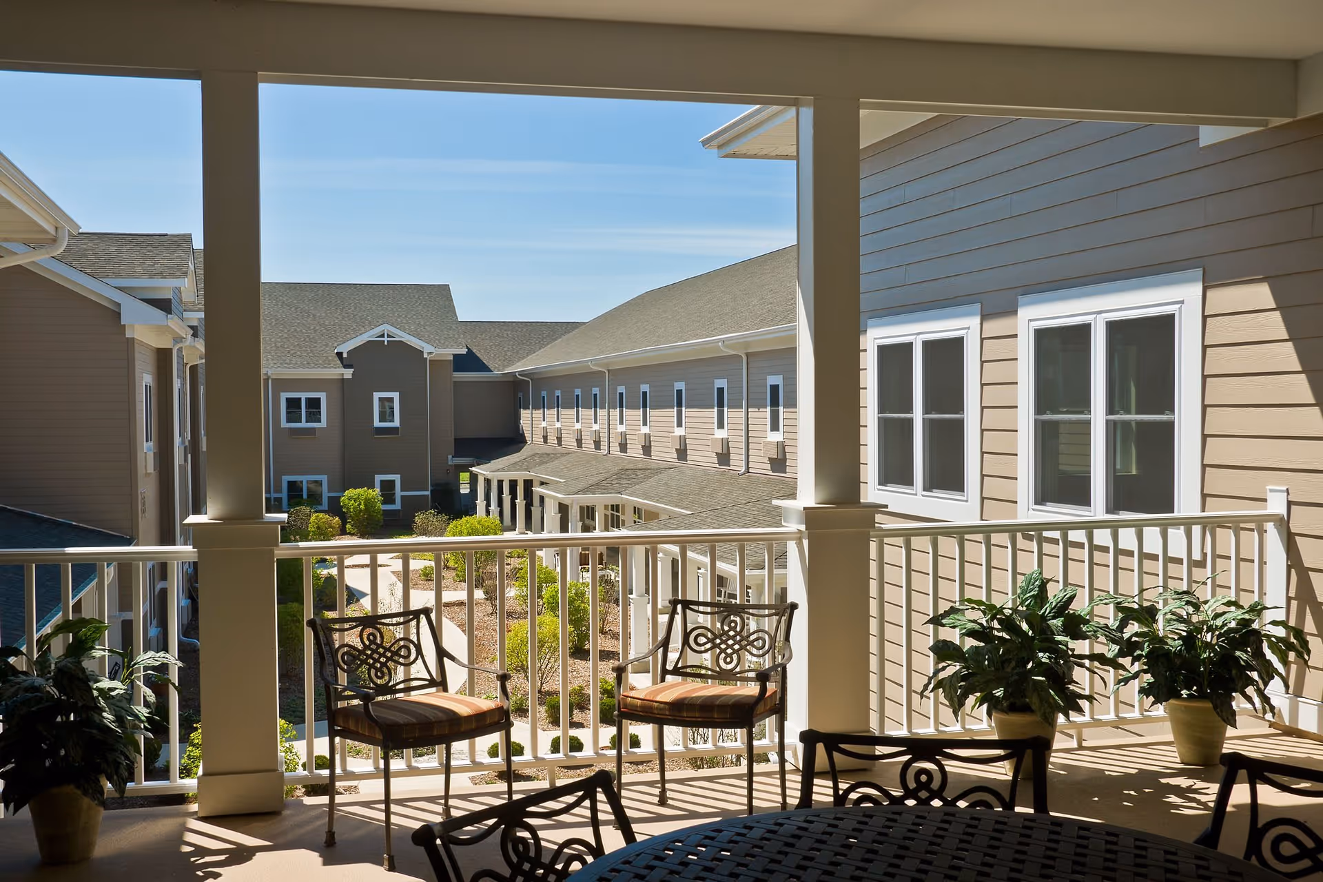 View from a covered balcony with metal chairs and potted plants overlooking a courtyard and surrounding buildings under a clear blue sky.