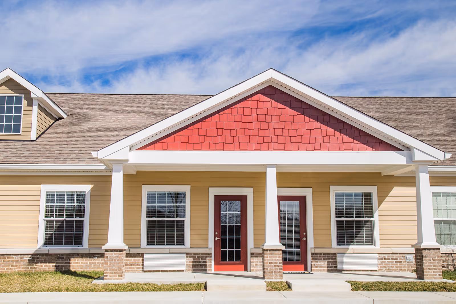 Front facade of a single-story building showing two red entry doors flanked by columns and windows beneath a red gable.