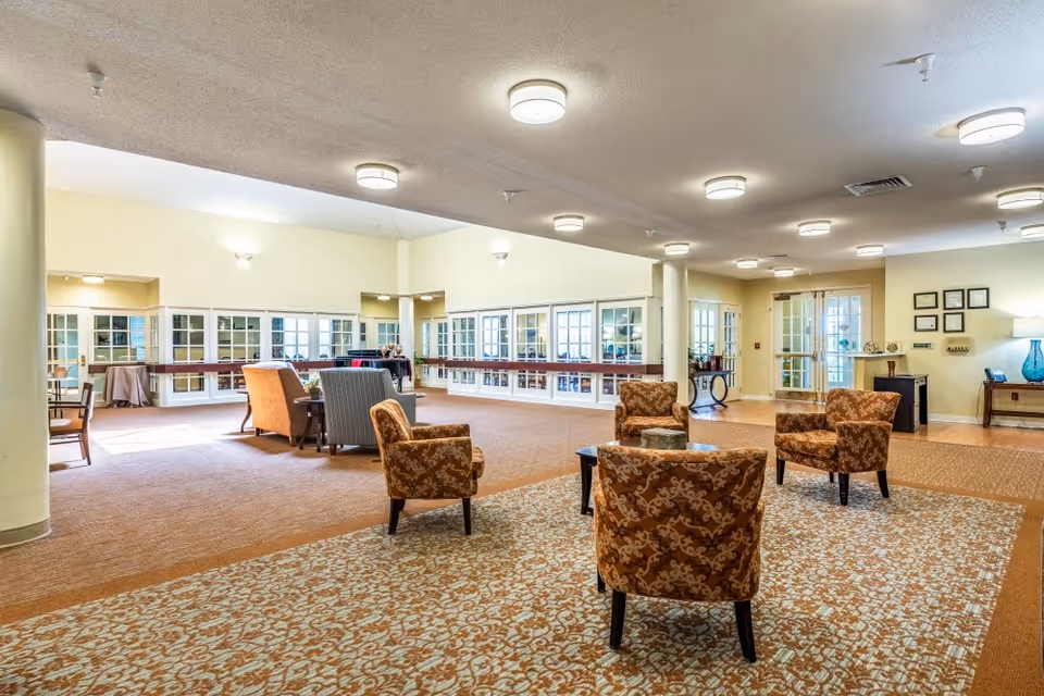 A spacious and well-lit common area in a senior living facility with patterned carpet and several upholstered armchairs arranged around small tables. The room features large windows with white frames, beige walls, and multiple ceiling lights. There are columns and a side table with a lamp and decorative items near the entrance.