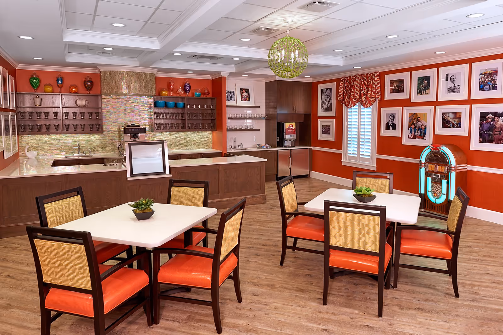 A bright and colorful dining area with two white tables, each surrounded by four chairs with orange seats and patterned backs. The room features a wooden floor, orange walls adorned with framed photos, a jukebox in the corner, and a kitchen counter with shelves holding glassware and colorful decorative jars. A green chandelier hangs from the ceiling.