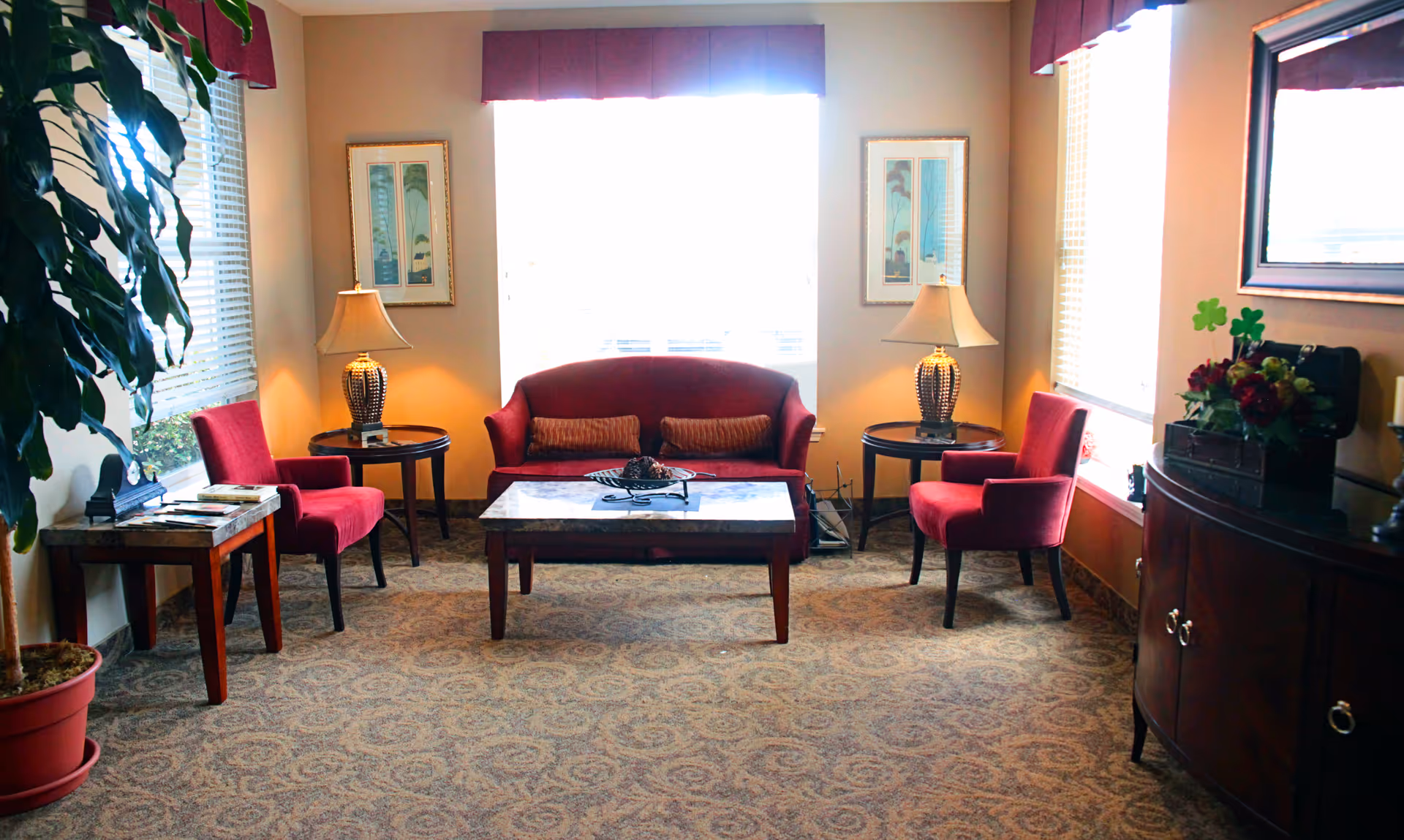 A cozy living room area with a red sofa, two red chairs, a coffee table, two side tables with lamps, framed artwork on the walls, a large window with blinds, a potted plant, and a wooden cabinet with decorative items.