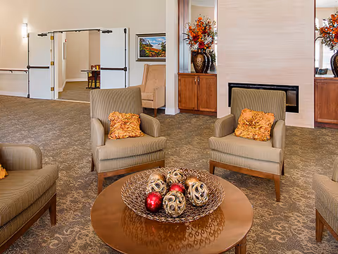 A cozy assisted-living lounge with upholstered armchairs arranged around a wooden coffee table topped with decorative orbs, floral arrangements and double doors in the background.