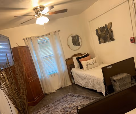 A cozy bedroom with a single bed featuring a white comforter and a decorative pillow with the word 'grateful'. The room has a ceiling fan with a light, a wooden armoire, a round mirror on the wall, decorative leaf wall art, a window with white curtains, and a patterned rug on the floor.