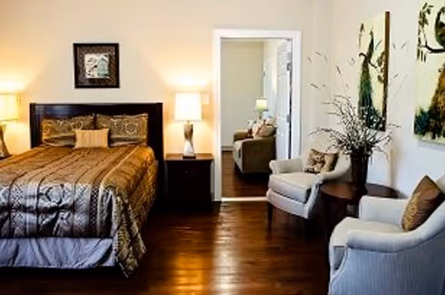 A cozy bedroom with a large bed covered in a patterned brown comforter and multiple pillows. There are two bedside tables with lamps on each side of the bed. To the right, there are two upholstered armchairs with cushions and a round wooden table between them holding a vase with decorative branches. The room has wooden flooring and light-colored walls adorned with two large paintings. A doorway leads to another room with a sofa and a lamp visible.