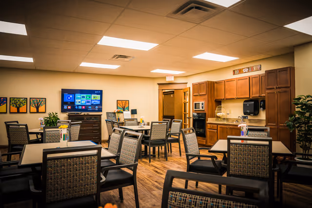 Well-lit common dining/activity room with multiple tables and chairs, a wall-mounted TV, and a kitchenette with wooden cabinets.