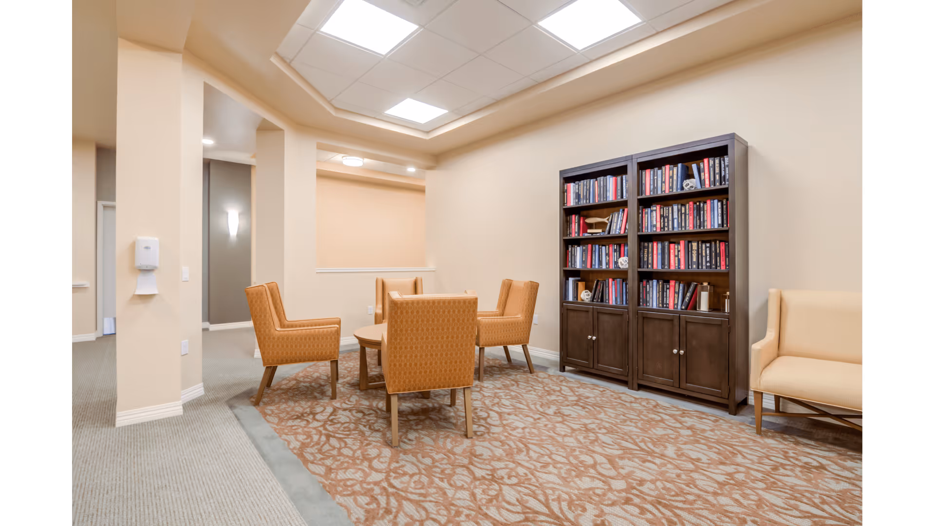 A cozy interior common area with a round wooden table surrounded by four orange upholstered chairs on a patterned carpet. There is a dark wooden bookshelf filled with books against the wall and a beige armchair to the right. The walls are painted light beige and the ceiling has recessed lighting.