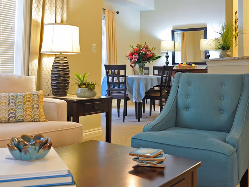 Bright furnished living room opening to a dining area with a teal armchair, sofa, coffee table, side table with a lamp, and a dining table topped with flowers.
