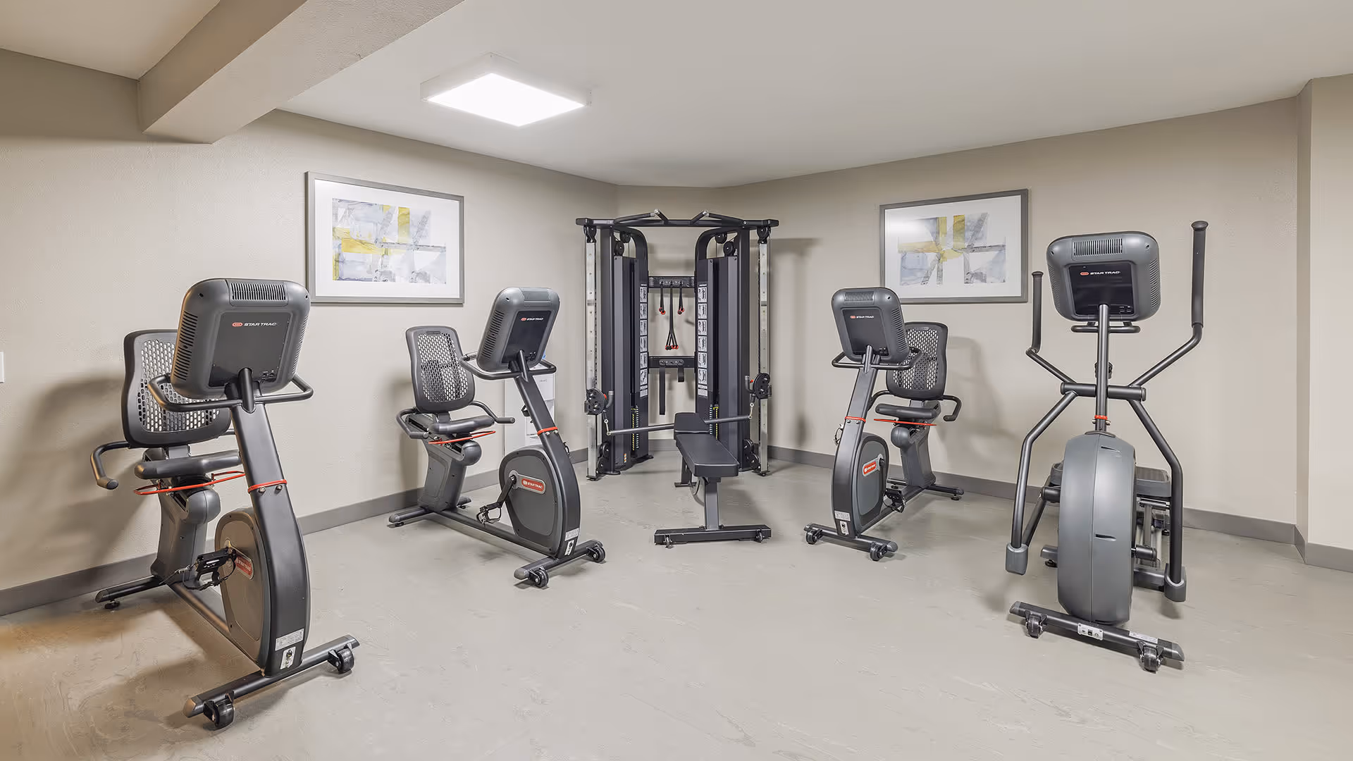A small fitness room with four exercise machines including two recumbent bikes, one elliptical trainer, and a multi-functional weight machine. The room has light gray walls, two abstract paintings, and a bright ceiling light.