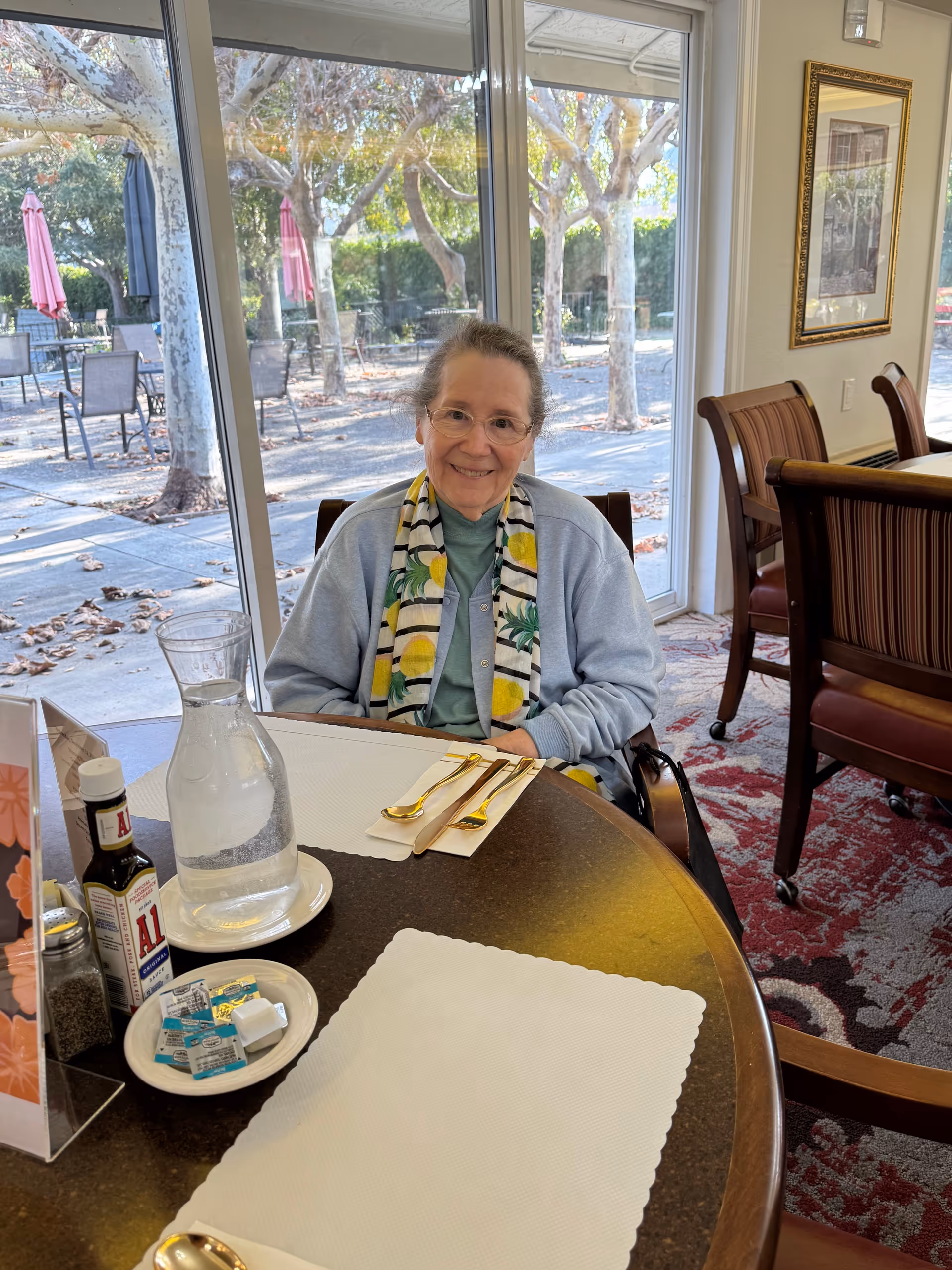 An elderly woman with glasses and a light blue cardigan sits at a dining table inside a retirement community dining room. She is smiling and wearing a scarf with a lemon pattern. On the table are a glass pitcher of water, a bottle of A1 sauce, salt and pepper shakers, butter packets, and gold-colored cutlery on white placemats. Behind her, large windows show an outdoor patio area with trees and tables with umbrellas.