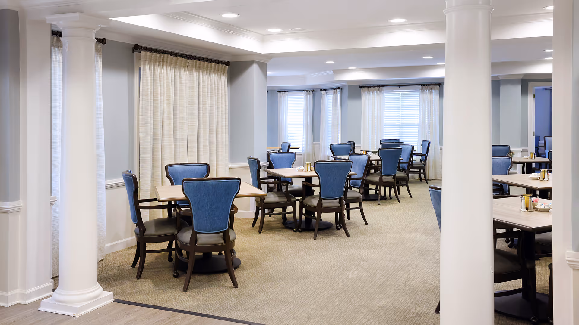 A bright dining room with multiple square wooden tables surrounded by blue upholstered chairs. The room features large windows with white curtains, white columns, and soft beige carpeting. The space is well-lit with recessed ceiling lights.