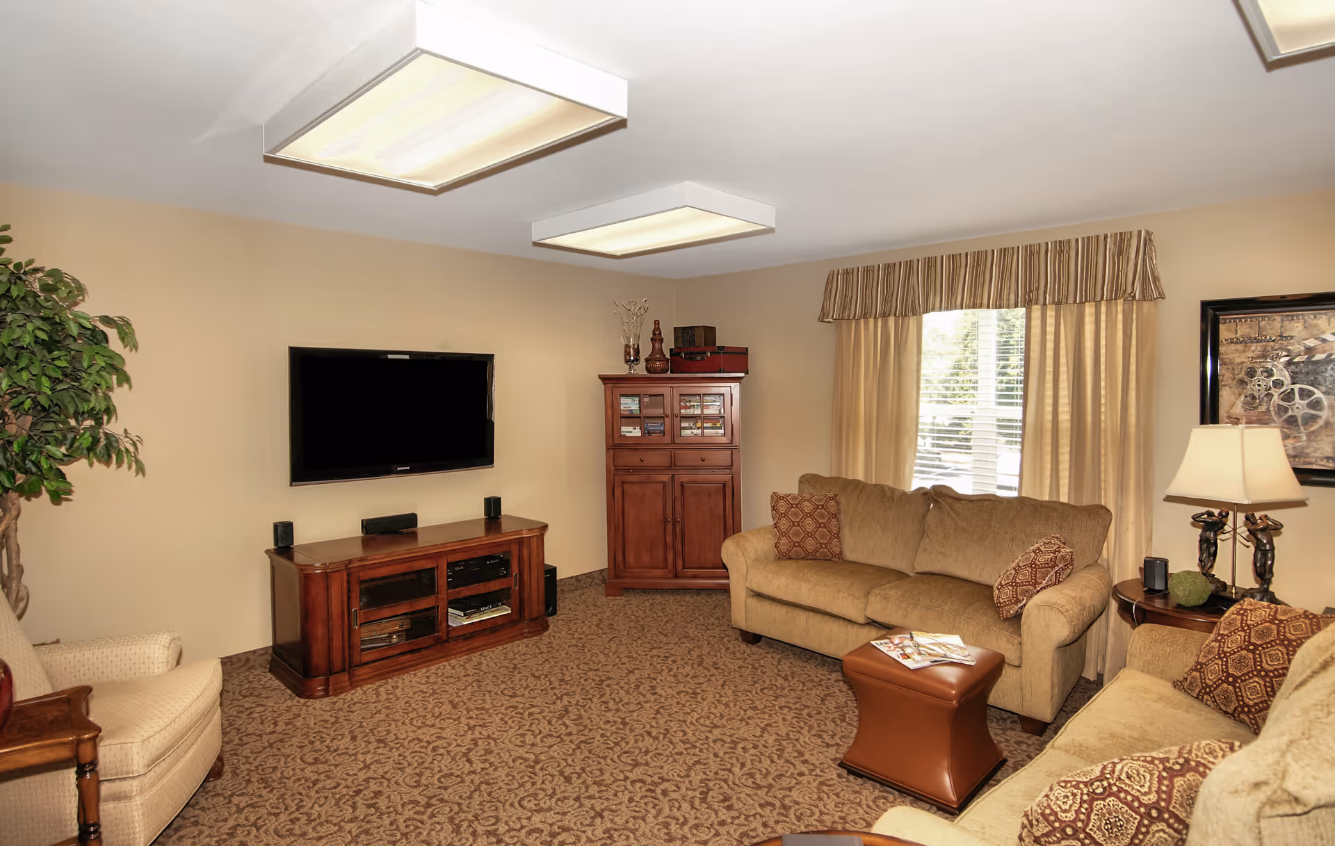 A cozy living room with beige walls and carpet, featuring two beige sofas with patterned cushions, a beige armchair, a wooden TV stand with a flat-screen TV mounted on the wall, a wooden cabinet with decorative items, a window with striped valance and curtains, a side table with a lamp, and a potted plant in the corner.
