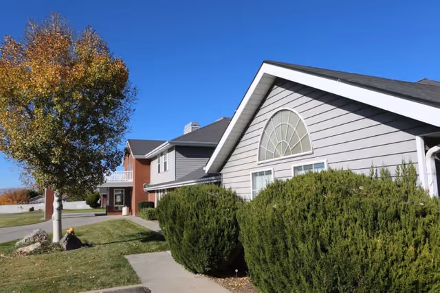 Exterior view of a senior living facility building with gray siding and a large arched window. There are green bushes along the sidewalk and a tree with autumn-colored leaves on the left side under a clear blue sky.