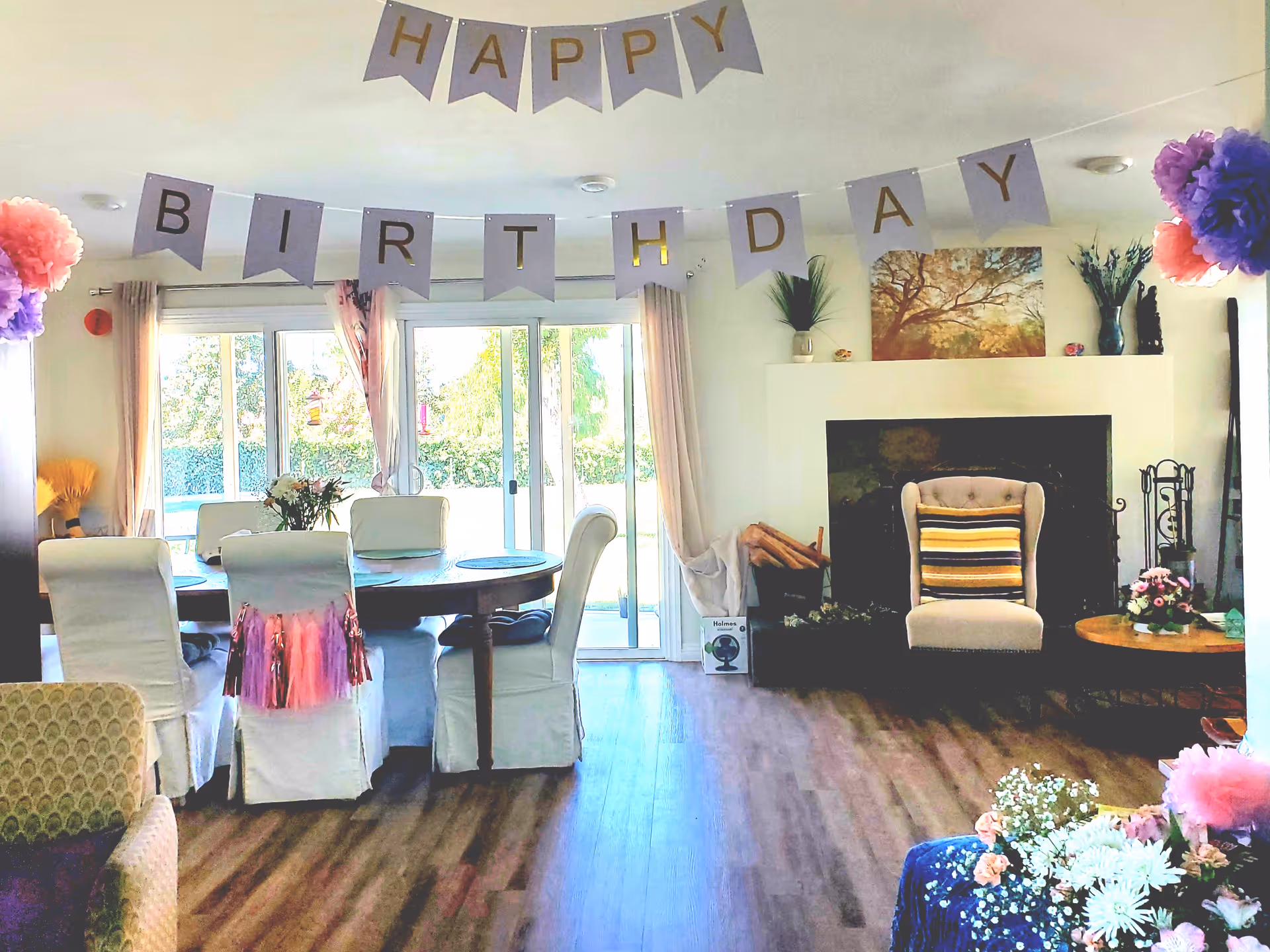 Bright communal dining and living area decorated with a "Happy Birthday" banner, a round dining table with white-covered chairs, and a fireplace with an armchair.