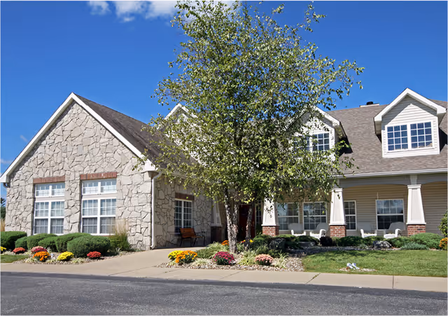 Exterior view of a senior living facility building named Riverbend with stone and siding facade, a tree in front, landscaped bushes and flowers, and a clear blue sky.