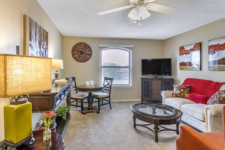 Bright furnished living room with a sofa, coffee table, TV, small round dining table, and decorative lamps and artwork.