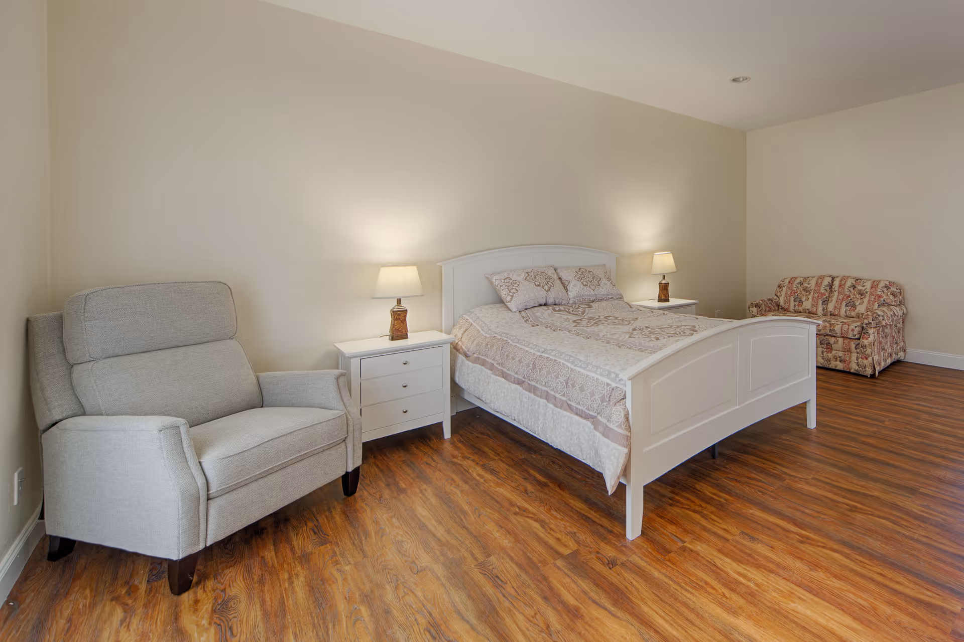 A bright bedroom with a white bed flanked by nightstands and lamps, a gray armchair to the left and a floral loveseat in the back on wood-look flooring.